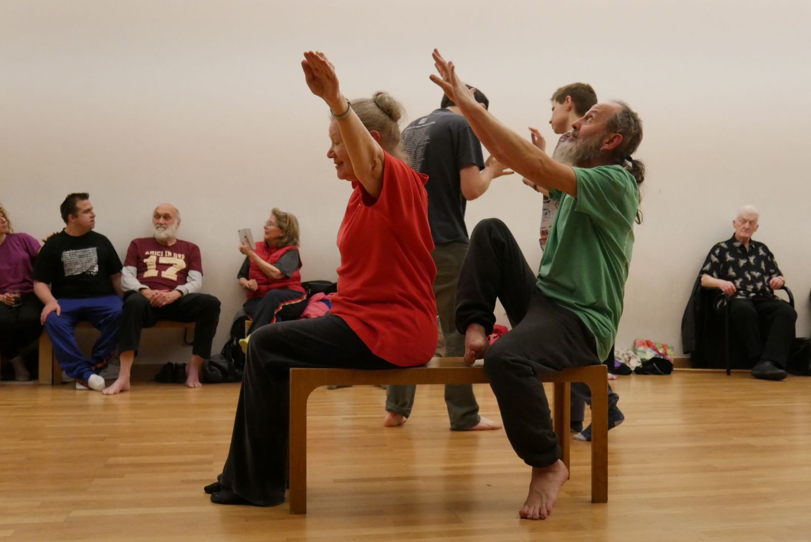 Two dancers at London’s Amici Dance Theatre Company sit on a bench. One woman reaches her arms to the sky and behind her a man spirals to reach toward her, head tipped up. Around them people sit and watch, and behind them two more dancers are touching hands, gently.